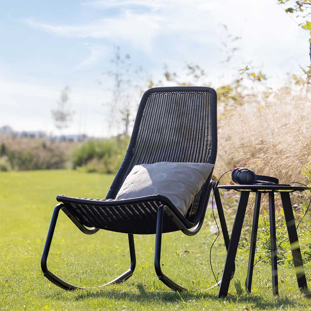 Skandi Gartenschaukelstuhl aus schwarzem Kunstrattan in grüner Outdoor-Umgebung – Vurgos